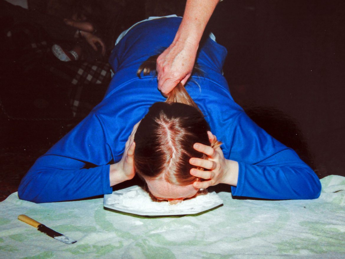 Person with long hair being held out the way as they put their face into a plate of flour to retrive a coin with their teeth.